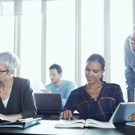 three students in an academic setting with computers and book where the elderly teacher is helping one of the female students