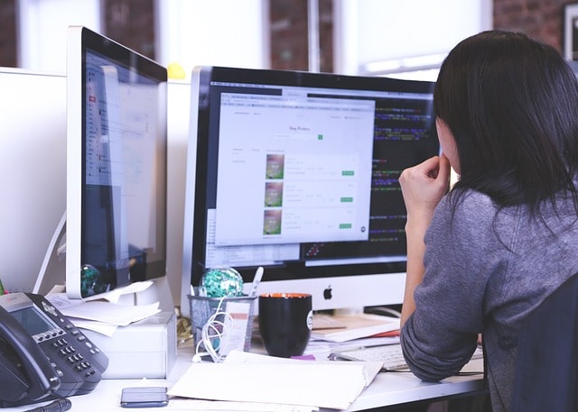 dark haired woman at computer with a phone and coffee and many documents around